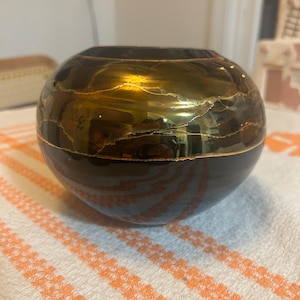 May include: A round, dark brown decorative bowl with a glossy finish. The bowl features gold-colored, abstract designs across the top half. The bowl is sitting on a white and orange striped cloth.