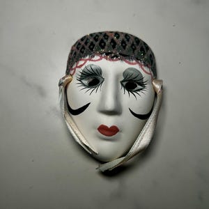 May include: A white decorative face mask featuring painted black and gray eye makeup with long lashes, red lips, and black curved cheek accents. The forehead area has a dark gray diamond pattern with pink trim, and light-colored ribbons are attached.