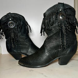 May include: A pair of black leather boots featuring a mid-calf height, a pointed toe, and a stacked heel. The upper shaft is decorated with black leather fringe, silver-toned metal studs, and a circular silver-toned concho ornament on each side.