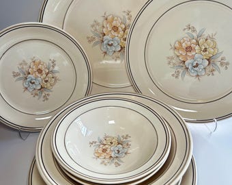 May include: A set of cream-colored ceramic dinnerware featuring a central floral motif of light blue, yellow, and peach-toned flowers. Each piece is accented with thin, dark brown circular bands along the rim, creating a cohesive look.