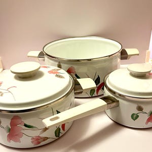 May include: A set of three white enamel cookware pieces featuring a floral pattern with pink flowers and green leaves. The set includes a large pot with two side handles, a medium pot with a lid, and a small saucepan with a long handle.