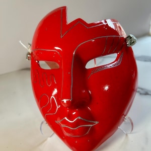 May include: A glossy red face mask featuring silver line art detailing across the forehead, cheeks, and lips. The mask has an asymmetrical cutout at the top and small silver bells attached to the sides near the eye openings.