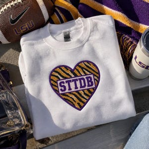 May include: A white crewneck sweatshirt featuring a central heart-shaped patch. The patch has a purple border and a yellow and black tiger stripe pattern. A white rectangular banner across the centre contains the purple text STTDB.