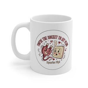 Pode incluir: Caneca de café de cerâmica branca com uma ilustração de desenho animado de um plugue e tomada com o texto "You're the Socket to My Plug" e "Sparks Fly!". O design inclui corações e estrelas.