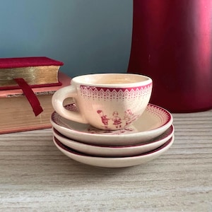 May include: A small, cream-colored teacup and saucer set with a burgundy floral design. The teacup is stacked on top of two matching saucers. The set has a vintage aesthetic, with a decorative border and illustrations of children.