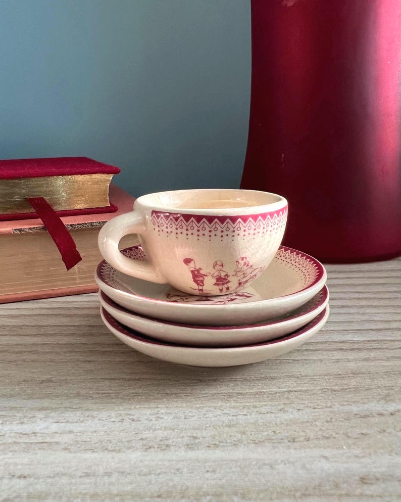May include: A small, cream-colored teacup and saucer set with a burgundy floral design. The teacup is stacked on top of two matching saucers. The set has a vintage aesthetic, with a decorative border and illustrations of children.
