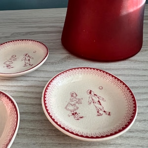 May include: Small, off-white ceramic plates with a red border and a decorative pattern. Each plate features a sepia-toned illustration of two children playing. A tall, red glass vase is in the background.
