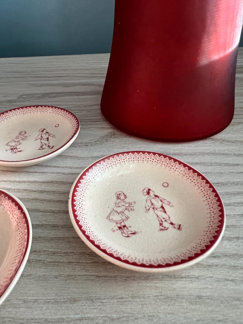 May include: Small, off-white ceramic plates with a red border and a decorative pattern. Each plate features a sepia-toned illustration of two children playing. A tall, red glass vase is in the background.
