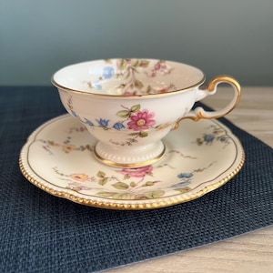 May include: A white ceramic mug with a black handle and the text "But First, Coffee" in a bold, black font. The mug is filled with a dark liquid, likely coffee, and sits on a light-colored surface.