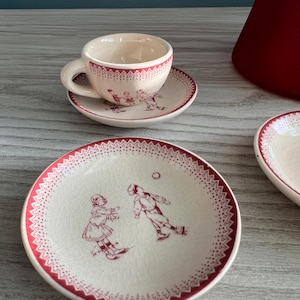 May include: A vintage-style teacup and saucer set with a cream-colored base and red trim. The set features a playful illustration of children playing, with a dotted border. The cup and saucer are small, likely for espresso or a demitasse.