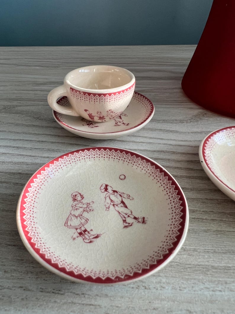 May include: A vintage-style teacup and saucer set with a cream-colored base and red trim. The set features a playful illustration of children playing, with a dotted border. The cup and saucer are small, likely for espresso or a demitasse.