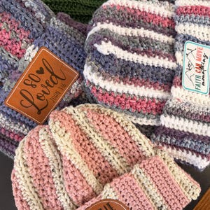 May include: A collection of four crocheted beanies in various colors including pink, cream, grey, and green. The beanies feature leather-like patches with text including "So Loved John 3:16", "Faith Can Move Mountains", and "Be The Light Matthew 5:14".