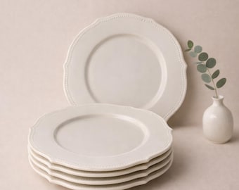May include: A stack of off-white ceramic plates with scalloped edges and a dotted border. Two additional plates are visible, along with a small white vase holding a sprig of greenery. The plates have a classic, elegant design.