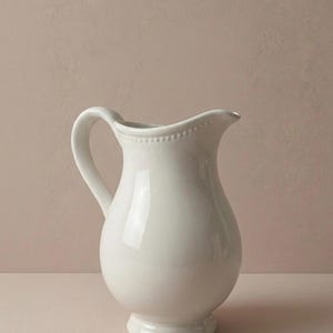 Puede incluir: Una jarra de cerámica blanca con un asa curva y un pico vertedor. La jarra tiene un cuerpo bulboso y una base pequeña y redonda. Una línea de pequeños puntos en relieve decora el borde. Ideal para servir bebidas.