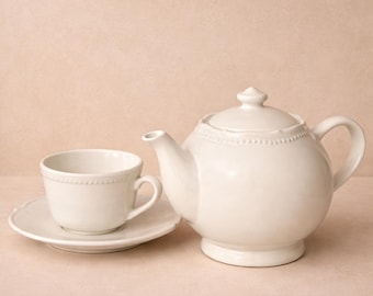 May include: A white ceramic tea set, including a teapot and a teacup with saucer. The teapot has a rounded shape with a lid and handle. The teacup and saucer are also white, with a delicate beaded edge. The set is designed for tea service.