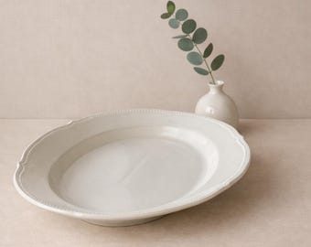 May include: An oval-shaped cream-colored ceramic serving platter with a beaded edge detail, accompanied by a small matching cream-colored ceramic bud vase containing a single sprig of green eucalyptus leaves.