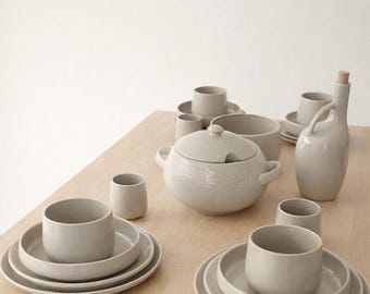 May include: A collection of neutral-toned ceramic tableware, including plates, bowls, cups, and a lidded soup tureen. A bottle with a wooden stopper and a pitcher are also present. The items are arranged on a light-colored wooden table.
