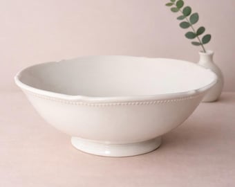 May include: A white ceramic serving bowl with a pedestal base. The bowl has a scalloped rim and a beaded detail around the edge. A small white vase with greenery is in the background. The bowl is suitable for serving salads or fruit.