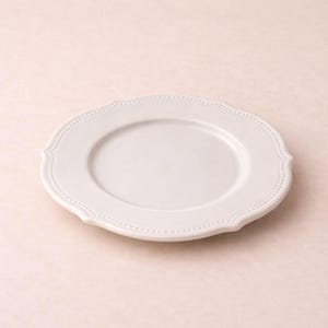 Può includere: Un piatto in ceramica bianca con bordo smerlato e bordo a pois. Il piatto ha un centro leggermente rialzato ed è progettato per servire cibo. Il piatto è di colore neutro e può essere utilizzato per varie occasioni.