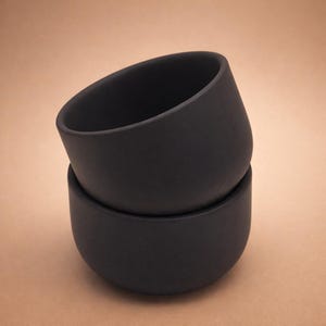 Könnte beinhalten: Zwei matte, schwarze Keramikschalen, die übereinander gestapelt sind. Die Schalen haben einen abgerundeten Boden und einen leicht abgewinkelten Rand. Die Schalen sind einfarbig dunkel und scheinen leer zu sein. Der Hintergrund ist hellbraun. Jede Schale hat einen Durchmesser von ca. 10 cm.