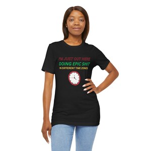 May include: Black t-shirt with the text "I'M JUST OUT HERE DOING EPIC SHIT IN DIFFERENT TIME ZONES" in red and green. A clock graphic with a red border is below the text. The shirt is worn by a person.