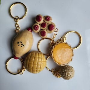 May include: A collection of five food-themed keychains featuring miniature charms shaped like a lemon, a round melon pan, a small tart with a yellow filling, a round sesame ball, and a flower-shaped cookie with red jam centres on gold rings.