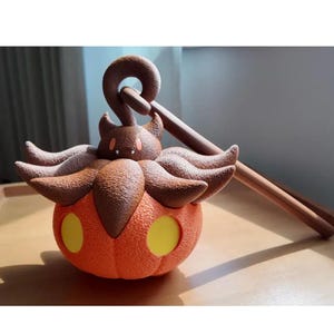 May include: An orange pumpkin-shaped lantern with yellow circular cutouts. The lantern has brown accents, including a handle and decorative elements. The design is reminiscent of a cartoon character. The lantern is resting on a wooden surface.