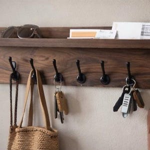May include: A dark wood wall-mounted shelf with six black hooks. Sunglasses and papers rest on the shelf. A woven bag, keys, and a leather pouch hang from the hooks. The shelf is mounted on a light-colored wall.