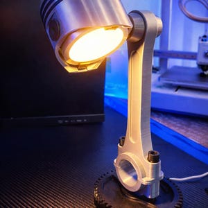 Könnte beinhalten: Eine einzigartige Schreibtischlampe aus recycelten Autoteilen. Die Lampe hat einen Kolbenkopf als Schirm, der ein warmes Licht ausstrahlt. Die Pleuelstange bildet den Stiel, und die Basis ist ein Zahnrad. Die Lampe ist silber und schwarz.