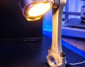 Op de afbeelding: Een unieke bureaulamp gemaakt van gerecyclede auto-onderdelen. De lamp heeft een zuigerkop als kap, die een warm licht uitstraalt. De drijfstang vormt de steel en de basis is een tandwiel. De lamp is zilver en zwart.