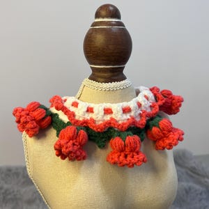 Puede incluir: Un collar de ganchillo hecho a mano con un diseño floral. El collar está adornado con flores rojas y hojas verdes, con un ribete estampado blanco y rojo. El collar se muestra en una cabeza de maniquí.