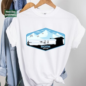 May include: White t-shirt featuring a blue and black graphic of the Hoth Echo Base from Star Wars. The design includes Tie fighters, AT-AT walkers, and the text "HOTH ECHO BASE" in white. The shirt is hanging on a wooden hanger.