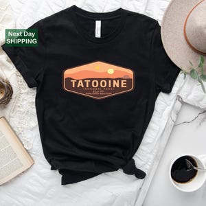 May include: Black t-shirt featuring a Tatooine National Park design. The graphic includes a desert landscape with two suns and the text "TATOOINE" above "NATIONAL PARK" and "ARKANIS SECTOR". The shirt is styled with a hat and a cup of coffee.
