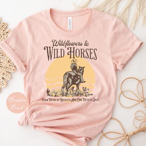 A pink t-shirt with a graphic design depicting a cowboy riding a horse, along with text that reads "Wildflowers & Wild Horses Four Fifties of Reckless and One Fiftieth of Jack".