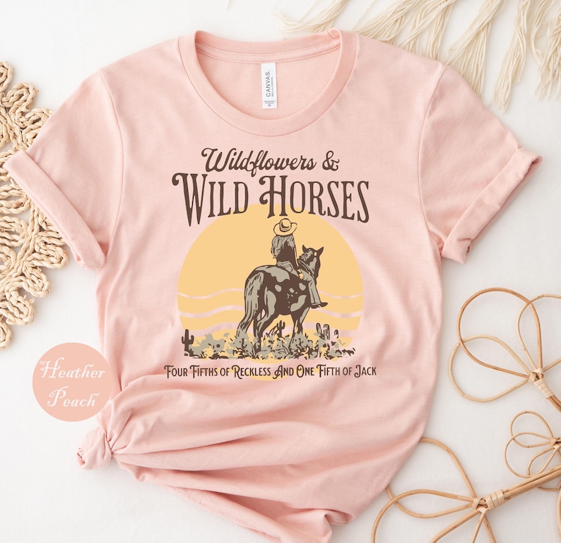 A pink t-shirt with a graphic design depicting a cowboy riding a horse, along with text that reads "Wildflowers & Wild Horses Four Fifties of Reckless and One Fiftieth of Jack".