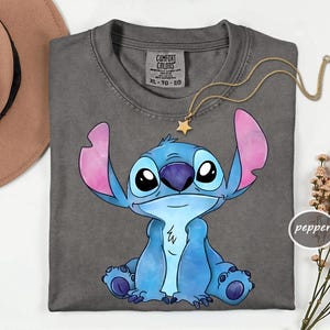 a gray t-shirt with a cartoon character design, specifically a blue and pink cartoon character resembling Lilo from the Lilo and Stitch movie franchise. The t-shirt is laid out on a white surface, with a hat and some flowers visible in the background.