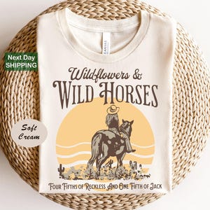 A white t-shirt with a graphic design depicting a cowboy riding a horse, with the text "Wildflowers & Wild Horses" and "Four Fifths of Reckless and One Fifth of Jack" printed on it.