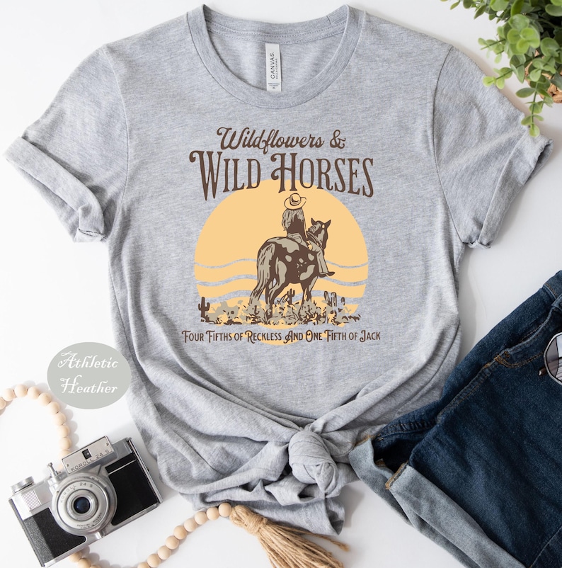 a gray t-shirt with a graphic design depicting a cowboy riding a horse, along with the text "Wildflowers & Wild Horses" and "Four Fives of Reckless and One Faith of Jack". The t-shirt is laid out on a white surface, accompanied by a pair of jeans, a camera, and a plant.