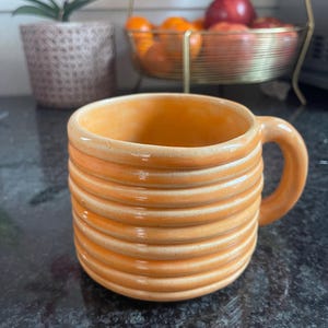 Puede incluir: Una taza de cerámica blanca con un asa negra y el texto "But First, Coffee" en negrita negra. La taza está sobre una superficie blanca. La taza mide aproximadamente 10 cm de alto.