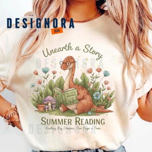 May include: Cream-colored t-shirt featuring a whimsical illustration of a brown dinosaur wearing glasses and reading a book titled "The Little Botanist." The design includes colorful flowers, mushrooms, and the text "Unearth a Story" and "Summer Reading."
