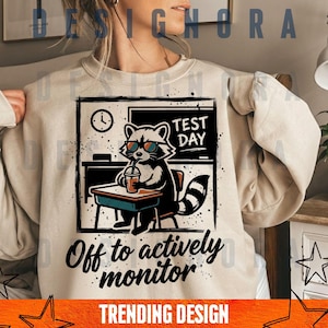 Könnte beinhalten: Beiges Sweatshirt mit einer Grafik eines Waschbären mit Sonnenbrille, der an einem Schreibtisch mit einem Getränk sitzt. Der Waschbär steht vor einer Tafel mit der Aufschrift "TEST DAY". Der Text "Off to actively monitor" steht unter dem Waschbären. Unten steht "TRENDING DESIGN".