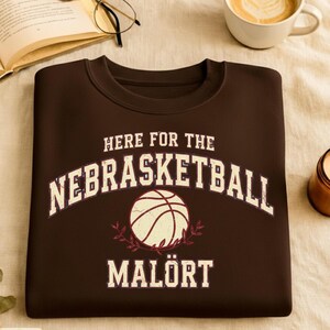 May include: A dark brown sweatshirt featuring the text "HERE FOR THE NEBRASKETBALL MALÖRT" in white, with a basketball graphic. The sweatshirt is on a beige surface, with a coffee cup and open book in the background.