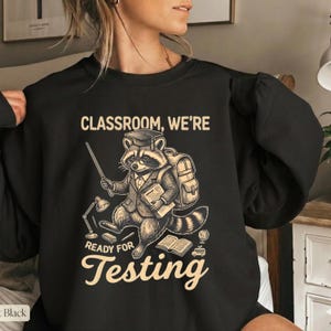 Könnte beinhalten: Schwarzes Sweatshirt mit einer Illustration eines Waschbären, der einen Abschluss-Hut, einen Anzug und einen Rucksack trägt. Der Waschbär hält einen Zeigestab und Bücher, mit dem Text "CLASSROOM, WE'RE READY FOR TESTING". Eine Lampe, ein Globus und ein Schulbus sind ebenfalls abgebildet.