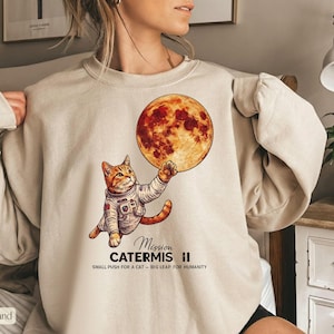 Puede incluir: Sudadera beige con un gato de dibujos animados con un traje espacial que alcanza una gran luna de color rojo anaranjado. El texto "Mission Catermis II" y "Small push for a cat - Big leap for humanity" está impreso debajo.