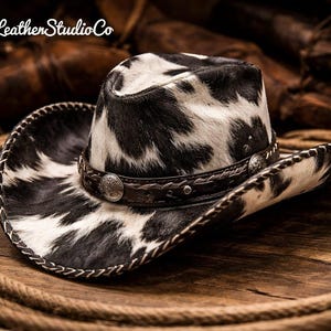 May include: Cowboy hat with a black and white spotted pattern. The hat has a brown leather band with silver-toned embellishments and a stitched brim. The hat is resting on a wooden surface with a rope.