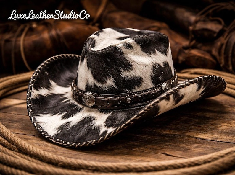 May include: Cowboy hat with a black and white spotted pattern. The hat has a brown leather band with silver-toned embellishments and a stitched brim. The hat is resting on a wooden surface with a rope.