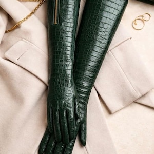 Könnte beinhalten: Ein Paar lange, dunkelgrüne Lederhandschuhe mit Krokodiltextur. Die Handschuhe haben einen goldenen Reißverschluss an der Seite und liegen auf einem beigen Mantel. Auch Accessoires wie eine goldene Kette und Ringe sind zu sehen.