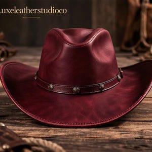 May include: A burgundy leather cowboy hat with a wide brim and a decorative band. The hat features a stitched design and is set on a wooden surface. The text "Luxeleatherstudioco" is visible in the upper left corner.