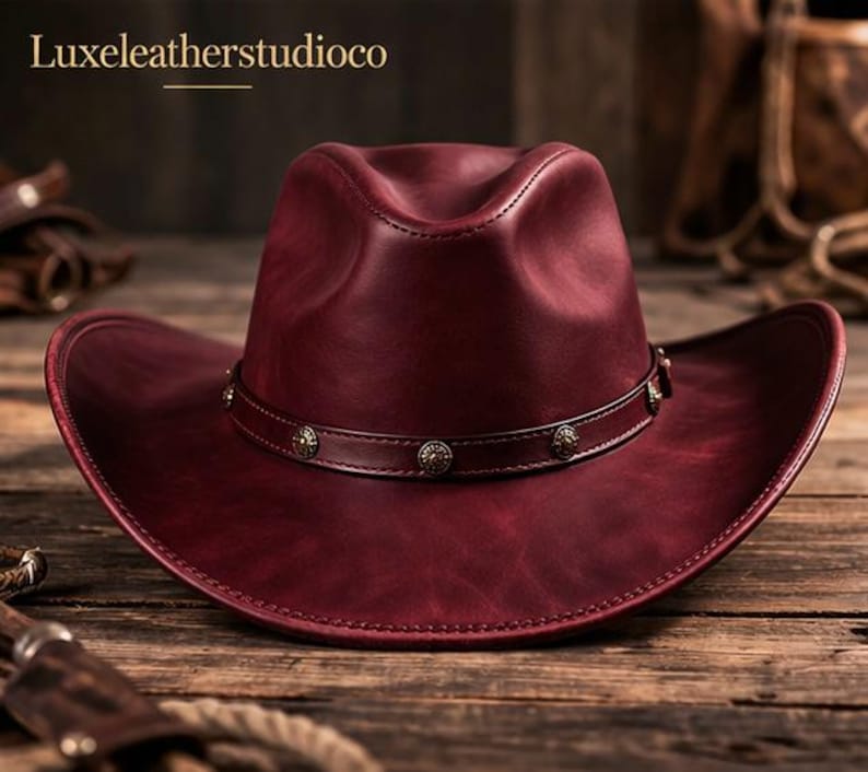 May include: A burgundy leather cowboy hat with a wide brim and a decorative band. The hat features a stitched design and is set on a wooden surface. The text "Luxeleatherstudioco" is visible in the upper left corner.
