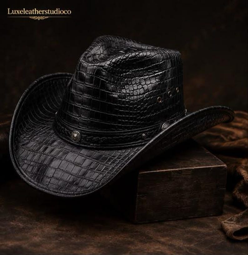 K&ouml;nnte beinhalten: Ein schwarzer Cowboyhut mit einer strukturierten Krokodilleder-Optik. Der Hut verf&uuml;gt &uuml;ber ein dekoratives Band mit Metallnieten und kleine, kreisf&ouml;rmige Bel&uuml;ftungs&ouml;sen an den Seiten der Krone. Die Krempe ist an beiden Seiten nach oben gebogen.